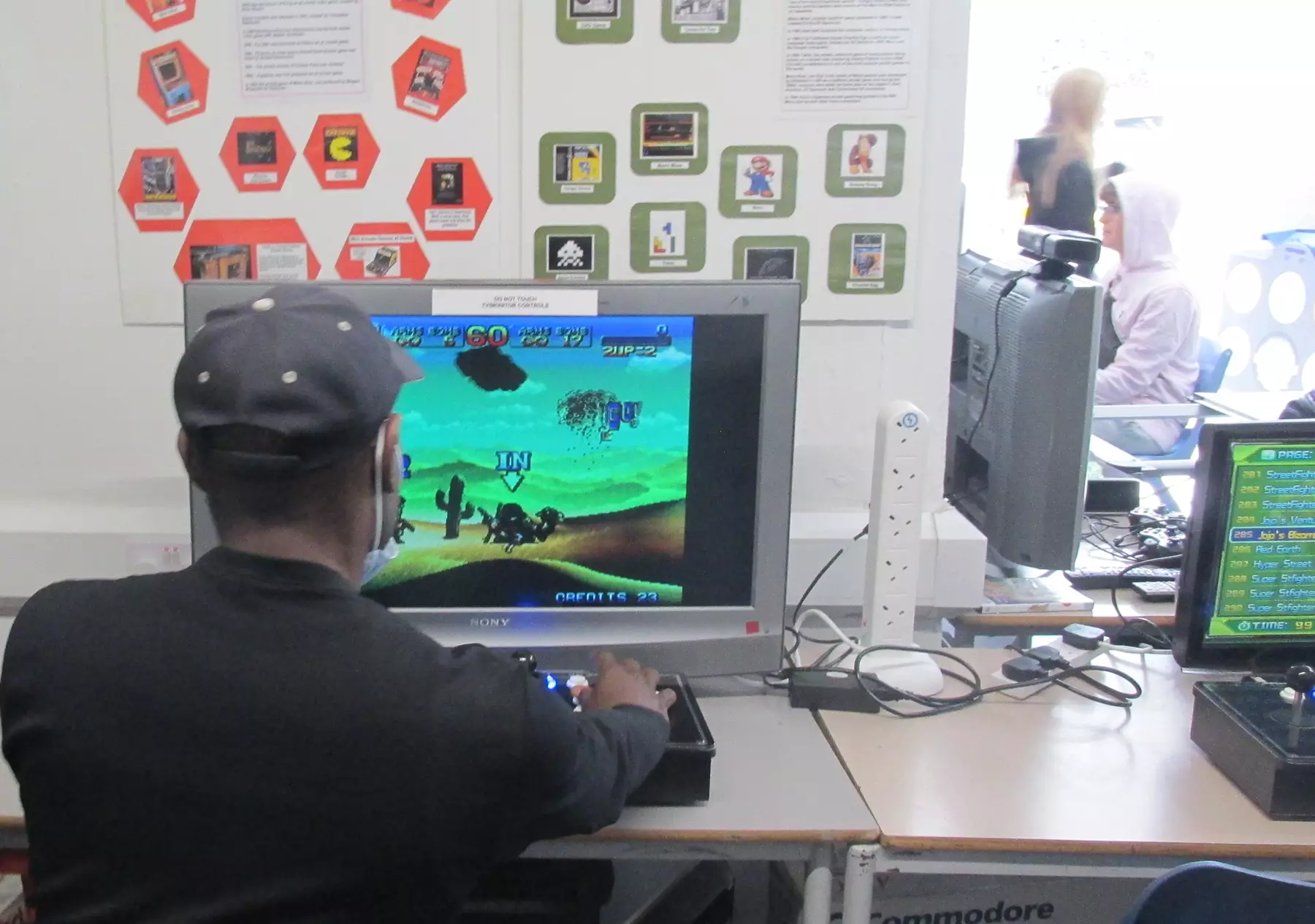 Man sat at computer screen playing game in front of wall of information. Two more computer screens can be seen and people in background